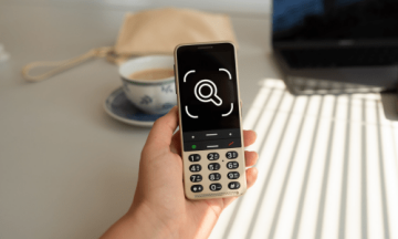 A person holding a BlindShell phone in their hand. The phone's display shows a symbol representing observo, consisting of magnifier inside the camera