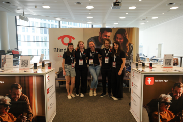 The BlindShell team standing together at the BlindShell booth during SightCity 2026 in Frankfurt. Five team members wearing event badges smile and pose in front of a large BlindShell Classic 3 backdrop. The booth features product displays, QR codes, and information about BlindShell features such as Tandem and Observo. The bright exhibition space is ready to welcome visitors interested in accessible technology for blind and visually impaired users.
