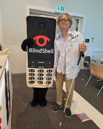 One of our lucky BlindShell Classic 3 winners poses proudly next to the new BlindShell mascot. Wearing sunglasses, a light jacket, and holding a white cane, the winner stands smiling beside the mascot. Together, they capture a heartwarming SightCity 2026 moment filled with excitement, gratitude, and the sense of community that makes these events so special.