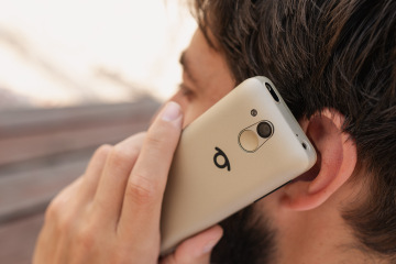Een man houdt een BlindShell Classic 3 in de kleur Goudkleurig zand tegen zijn oor tijdens een telefoongesprek. Op de foto is de achterkant van de telefoon te zien, inclusief de camera, de SOS-knop en het BlindShell-logo.