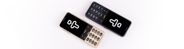 Twee BlindShell Classic 3-telefoons liggen diagonaal op een schone witte achtergrond. Eén telefoon heeft de kleur Goudkleurig zand en de andere Oceaanblauw. Op beide schermen staat het pictogram van de Tandem-app, gevormd als de letter T, die ook lijkt op een menselijke figuur. De afbeelding is helder, duidelijk en professioneel vormgegeven.