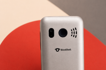 Close-up of the upper back of a silver BlindShell Classic Lite phone against a warm beige, red, and dark gray geometric background. The image highlights the round SOS button and BlindShell logo.