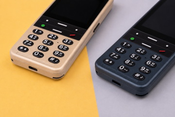 Close-up of two BlindShell Classic 3 phones in Golden Sand and Ocean Blue displayed side by side on a soft yellow and light gray background. The image highlights their large tactile keypads with clearly distinguishable buttons, call controls, speaker grilles, and charging ports.