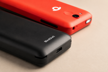 Close-up of two BlindShell Classic 2 phones overlapping on a warm beige background. A black BlindShell Classic 2 is shown in the foreground with the charging port and BlindShell logo visible, while a red BlindShell Classic 2 in the background shows its camera, SOS button, side controls, and BlindShell logo.