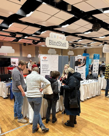 Bezoekers verzamelen zich rond de BlindShell-stand op de INSPO-conferentie voor ondersteunende technologie en luisteren naar productdemonstraties terwijl ze vragen stellen in een drukke beurshal.