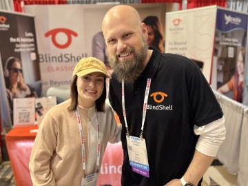 Molly Burke en Sam van The Blind Life staan dicht bij elkaar en glimlachen bij de BlindShell-stand tijdens een conferentie over ondersteunende technologie. Molly draagt een lichtgekleurde pet en een beige trui en heeft een conferentiebadge aan een keycord. Sam draagt een zwart BlindShell-poloshirt met het BlindShell-logo en een exhibitorbadge. Beiden zien er ontspannen en vriendelijk uit. Achter hen zijn BlindShell-banners en promotiemateriaal te zien.