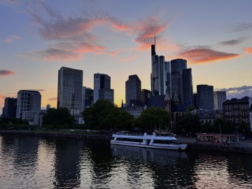 Frankfurt's skyline at sunset, with modern skyscrapers reflecting the last light of the day. The city's distinctive high rise buildings rise above the River Main, where boats are moored along the waterfront. Pink and orange clouds stretch across the evening sky, creating a picturesque view of Germany's financial capital, often nicknamed "Mainhattan.