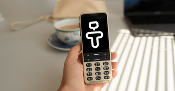 A hand holding Golden Sand BlindShell Classic 3 with a Tandem logo on the display. Behind the phone is a white and blue coffee mug. The whole image has vibrant colors